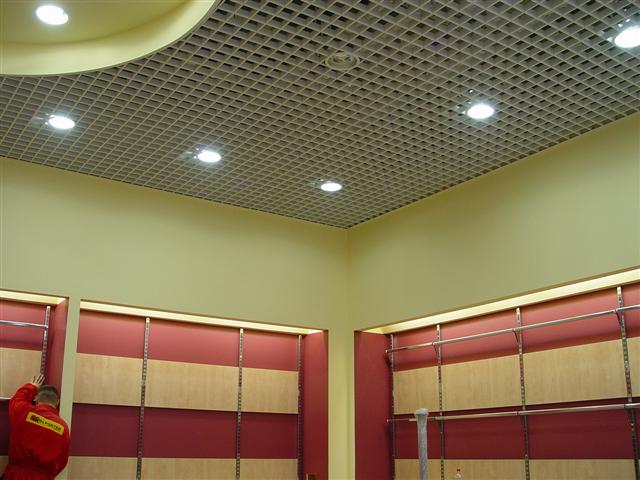 Kasetonowy sufit podwieszany z oświetleniem punktowym w lokalu komercyjnym, widoczny pracownik w czerwonym kombinezonie.